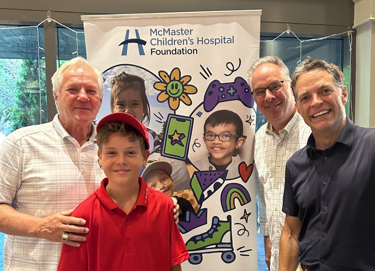 A group photo of four smiling men and a young boy standing in front of a McMaster Children’s Hospital Foundation banner.